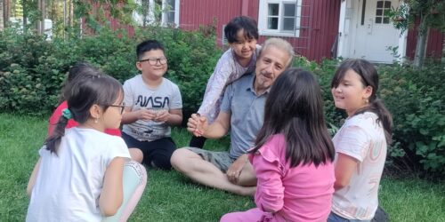 Oma kieli ja kulttuuri kuvakaruselli Aikuinen ja lapsia istuvat ringissä nurmikolla.